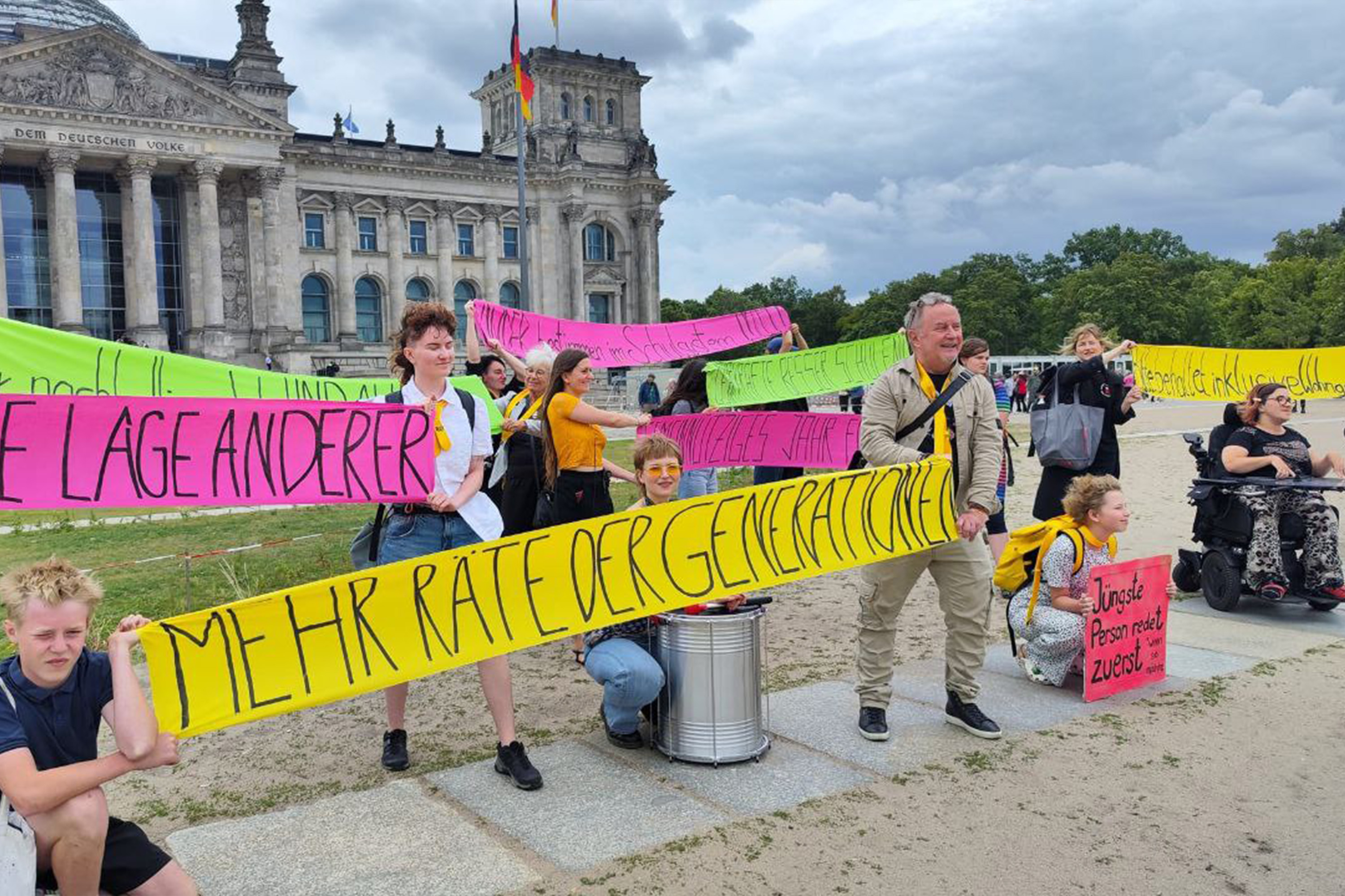 Foto vom „Rat der Generationen“ in Berlin. Einige Ratsmitglieder stehen mit bunten Bannern aus dem Theater vor dem deutschen Bundestag. Sie haben eine Trommel und das pinke Schild „Jüngste Person redet zuerst!“ dabei. Auf den Bannern ist zum Beispiel zu lesen: „Mehr Räte der Generationen“ oder „Lehrkräfte besser schulen“ oder „Gemeinnütziges Jahr“. Es weht starker Wind und es sind dunkle Wolken am Himmel zu sehen.